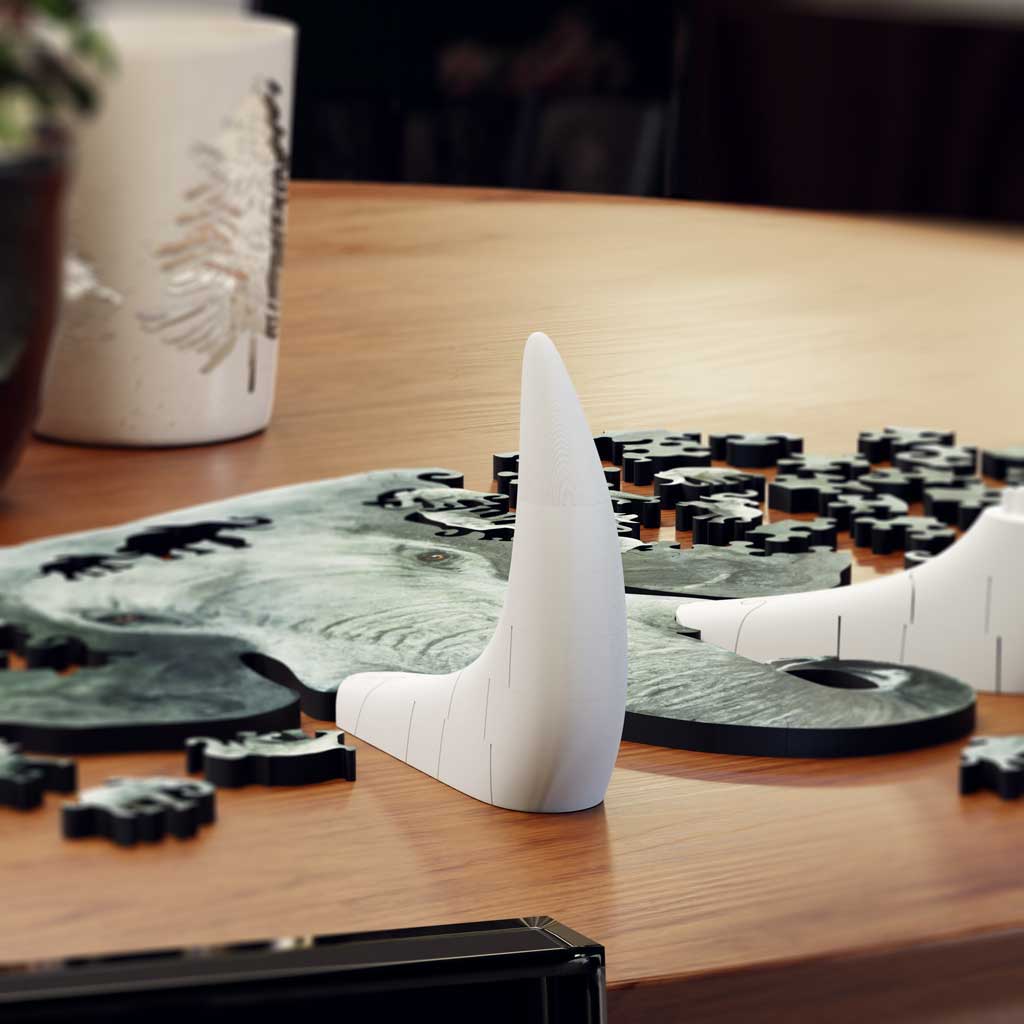 Lifestyle scene of the elephant puzzle in progress on a round wooden table, scattered pieces and tusk sections in front of candles and a small potted plant.