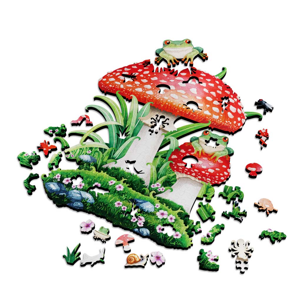 Wooden jigsaw puzzle of two red and white toadstool mushrooms with tree frogs perched on top, partially assembled and surrounded by whimsy-cut pieces shaped like snails, flies, beetles, flowers, and blades of grass.
