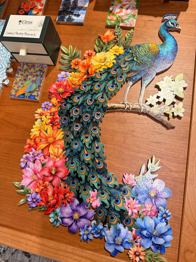 A colorful wooden jigsaw puzzle featuring a peacock with a long tail of flowers, displayed on a wooden tabletop next to its packaging box.