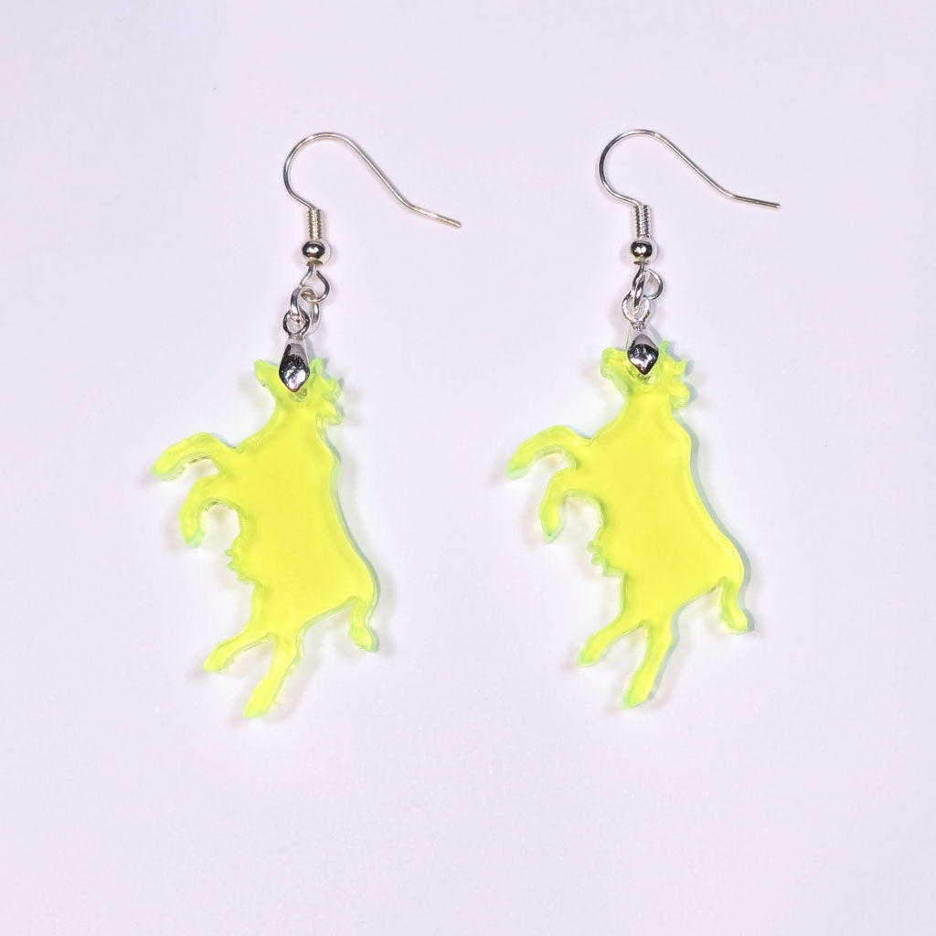 A pair of neon green translucent acrylic cow-shaped earrings with silver hooks, laid flat against a light lavender background. The cow silhouettes are slightly irregular with organic edges, and cast soft shadows beneath them.