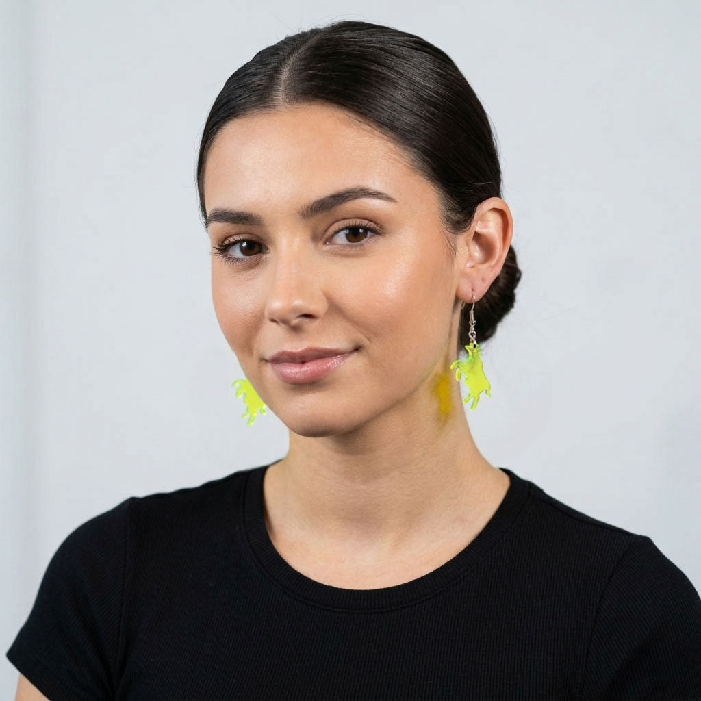 A woman with dark hair in a low bun, wearing a black fitted t-shirt, faces the camera with a slight smile. She wears a pair of neon green acrylic cow earrings, which are visible on both ears against a neutral gray background.